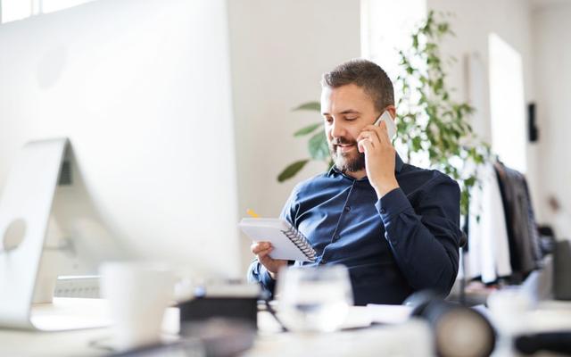 Arbeitslos melden: Mann sitzt an seinem Schreibtisch und telefoniert übers Handy mit dem Arbeitsamt, während er auf den Notizblock in seiner anderen Hand schaut.
