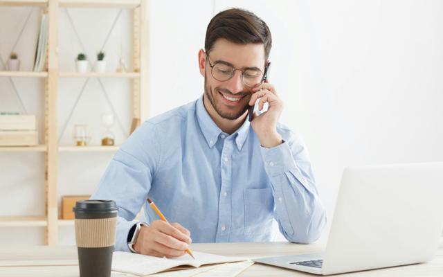 Junger Geschäftsmann sitzt vor seinem Laptop, telefoniert mit dem Handy und macht Notizen, während er sich zum Thema Aufstiegsfortbildung beraten lässt.