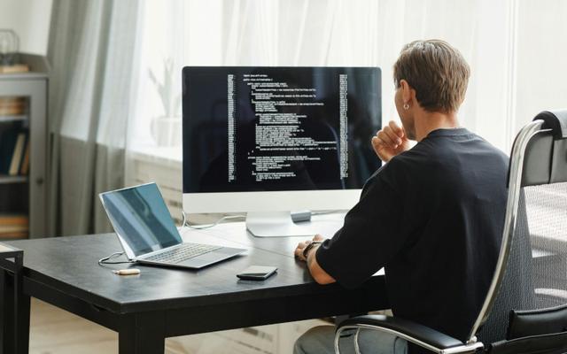 Ein junger JavaScript-Entwickler sitzt am Schreibtisch vor einem großen Bildschirm mit Quellcode und einem aufgeklappten Laptop.
