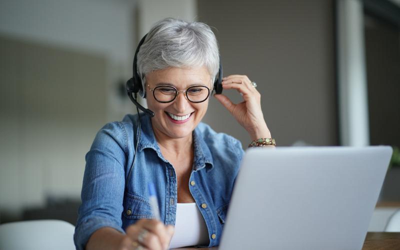 Mit 50 Jobwechsel: Frau über 50 mit kurzen grauen Haaren und Headset sitzt lächelnd und mit einem Stift in der Hand vor dem Laptop und folgt dem Unterricht.