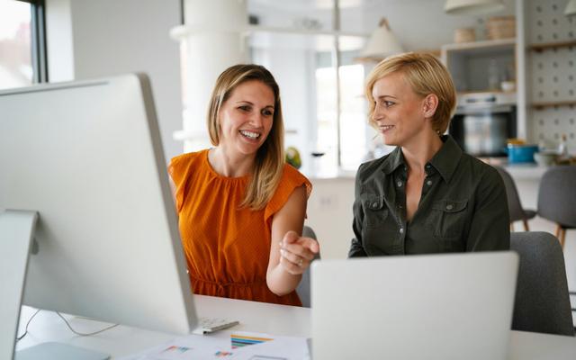 Mitarbeiterin im Büro erklärt ihrer kurzhaarigen Kollegin, die neben ihr am Schreibtisch sitzt, in einer SAP Schulung die Grundlagen des Systems.