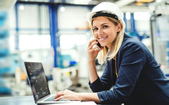 Nach erfolgreich bestandenem SPS Kurs arbeitet die blonde Frau jetzt als SPS-Technikerin - mit weißem Schutzhelm und Laptop - an in der Industrie. 