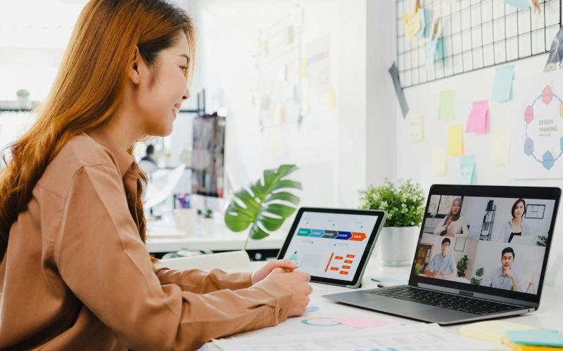 Junge Frau mit langen Haaren sitzt an ihrem Arbeitsplatz vor zwei Laptops und folgt online ihrer Teilzeit-Umschulung.