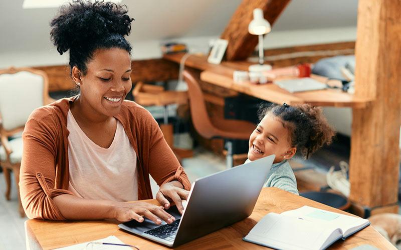 Frau sitzt an einem kleinen Wohnzimmertisch vor ihrem Laptop und nimmt an ihrer Teilzeit-Weiterbildung teil, während ihre kleine Tochter neben ihr steht.