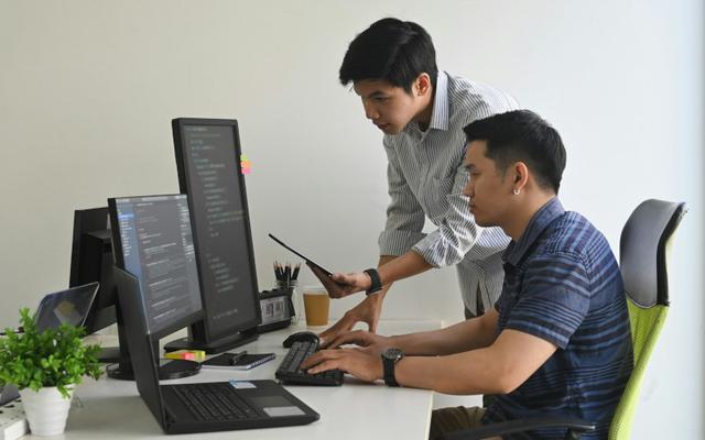 IT-Spezialist steht neben einem Umschüler, der an einem Schreibtisch mit zwei Monitoren sitzt, und gibt ihm praktische Tipps während seiner Umschulung Fachinformatik.