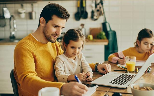 Vater sitzt mit seinen beiden Töchtern am Küchentisch und nimmt gleichzeitig an einer Weiterbildung Teilzeit teil.