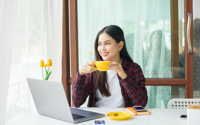 Lächelnde junge Frau sitzt mit Kaffeetasse auf der Terrasse vor ihrem Laptop und denkt über ihre Weiterbildungsmöglichkeiten nach.