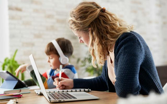 Junge Mutter sitzt am Laptop, lernt und bereitet ihren Wiedereinstieg nach Elternzeit vor, während hinter ihr ihr kleiner Sohn mit Kopfhörern sitzt.