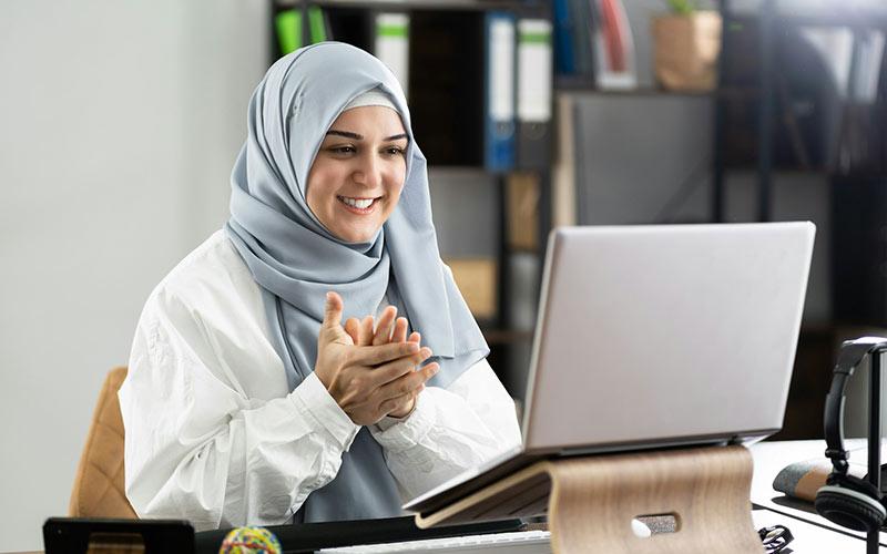 Junge Frau mit Kopftuch sitzt an einem modernen Arbeitsplatz vor einem aufgeklappten Laptop und erlernt begeistert die Ausbildungsbausteine & Qualifizierungsbausteine ihrer Teilqualifikation.