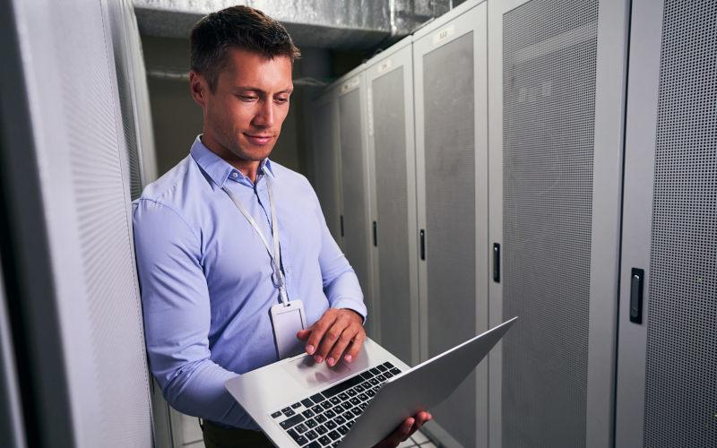 Cloud Computing: IT-Spezialist steht mit Laptop in der Hand im Serverraum und prüft das Netzwerk.