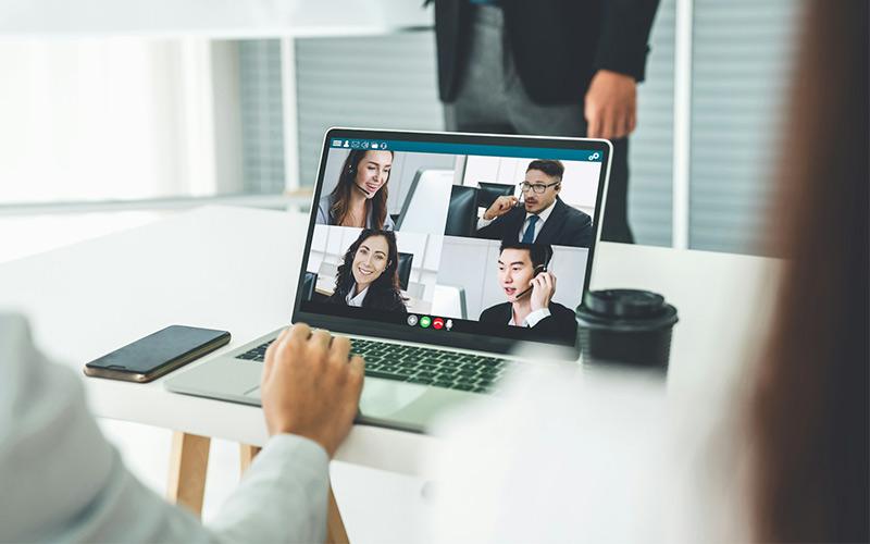 Digitalisierung im Arbeitsleben: Auf einem Laptop sind Kollegen in einer Videokonferenz zu sehen, die zusammen mit der Kollegin im Büro an der Präsentation des Chefs teilnehmen.