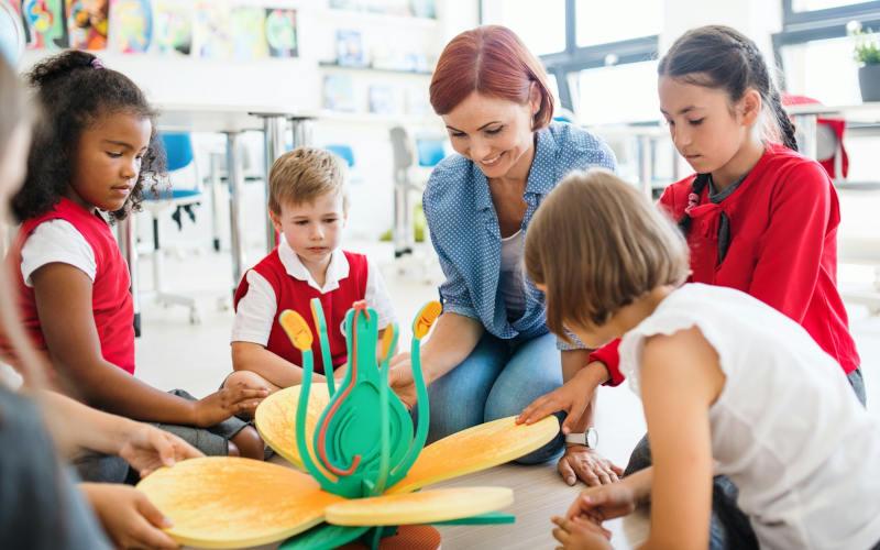 Rothaarige Erzieherin sitzt mit einer Gruppe von Kindergartenkindern um eine hölzerne herum und muss sich aufgrund des Fachkräftemangel Kita allein um sie kümmern.