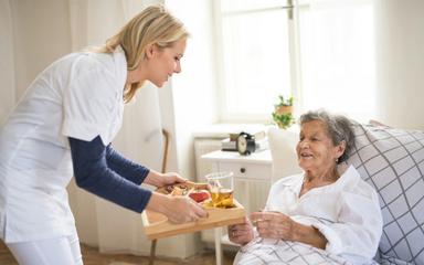 Fachkräftemangel Pflege: Junge Pflegerin bringt einer alten Frau ein Tablett mit Frühstück ans Bett.