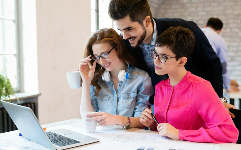 Zwei junge Frauen sitzen während ihrer Umschulung gemeinsam vor einem Laptop, während ihnen ein junger Dozent im Anzug über die Schulter schaut. 