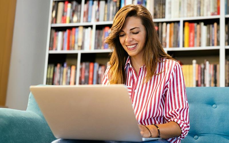 Der Frauenanteil bei Weiterbildungen ist hoch: Frau sitzt mit ihrem Laptop auf dem Sofa, hinter ihr ein breites Regal voller Bücher.