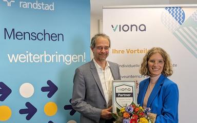 Dr. Christoph Kahlenberg (Manager Randstad Akademie & Arbeitsmarktprojekte) und Lea Tornow (Viona-Geschäftsführerin) stehen vor einem Randstad und einem Viona-Rollup und besiegeln mit einem Blumenstrauß und dem Viona-Partner-Zertifikat feierlich die Kooperation.