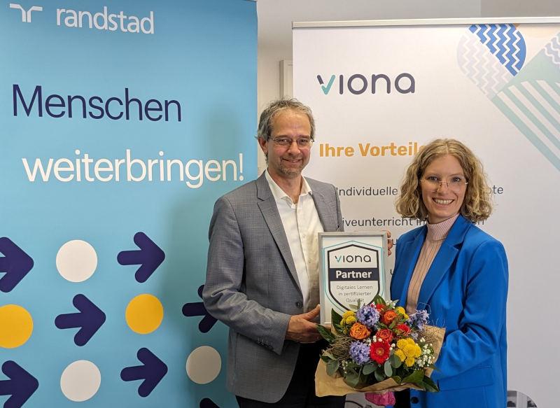 Dr. Christoph Kahlenberg (Manager Randstad Akademie & Arbeitsmarktprojekte) und Lea Tornow (Viona-Geschäftsführerin) stehen vor einem Randstad und einem Viona-Rollup und besiegeln mit einem Blumenstrauß und dem Viona-Partner-Zertifikat feierlich die Kooperation.