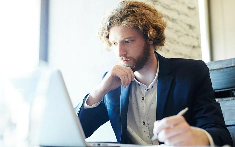 Bärtiger junger Mann mit Locken sitzt grübelnd und mit Stift in der Hand vor seinem Laptop und überlegt, was bei seiner Weiterbildung schiefgelaufen ist.