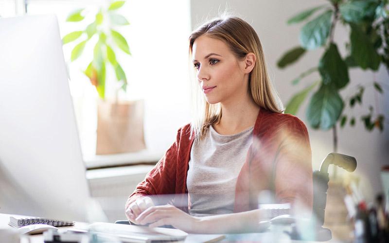 Junge Frau sitzt an ihrem Schreibtisch im Büro, von Pflanzen umgeben, und recherchiert an ihrem Computer Weiterbildungen für IT-Fachkräfte.