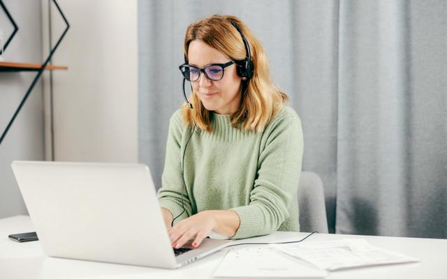 Blonde Frau mit Headset und Brille informiert sich an ihrem Laptop über Umschulungen durch das Arbeitsamt.