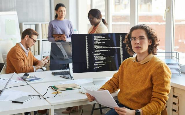 Future Skills: Ein junger IT-Entwickler vor seinem Bildschirm mit Quellcode schaut in die Kamera, während sich im Hintergrund drei Kolleg:innen unterhalten.