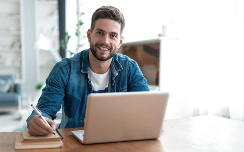 Lächelnder junger Mann sitzt am Schreibtisch vor seinem Laptop und macht Notizen während seiner Umschulung über das Arbeitsamt.